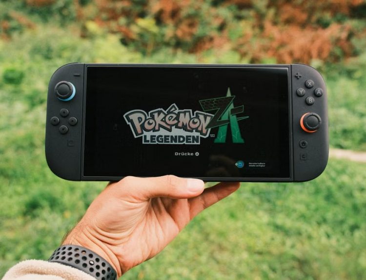 A hand holding a handheld gaming console displaying the Pokémon Legends game screen outdoors.