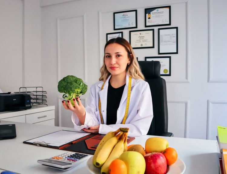 Female nutritionist in office holding broccoli, surrounded by fruits, promoting healthy lifestyle.