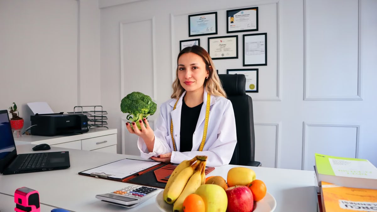 Female nutritionist in office holding broccoli, surrounded by fruits, promoting healthy lifestyle.